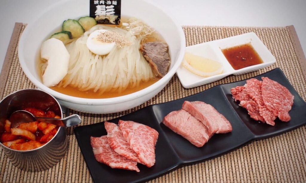Cold reimen soup with separate kimchi and raw meat to be barbecued.
